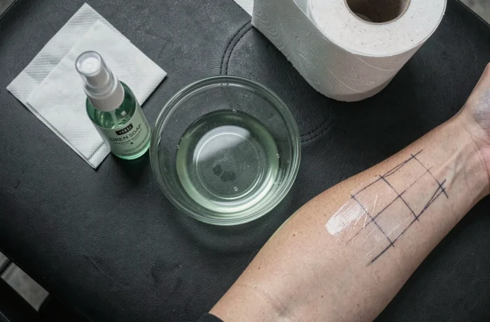 Vue en plongée de la station de préparation d'un tatoueur avec savon vert, compresses stériles et avant-bras montrant l'effacement partiel d'un stencil de tatouage